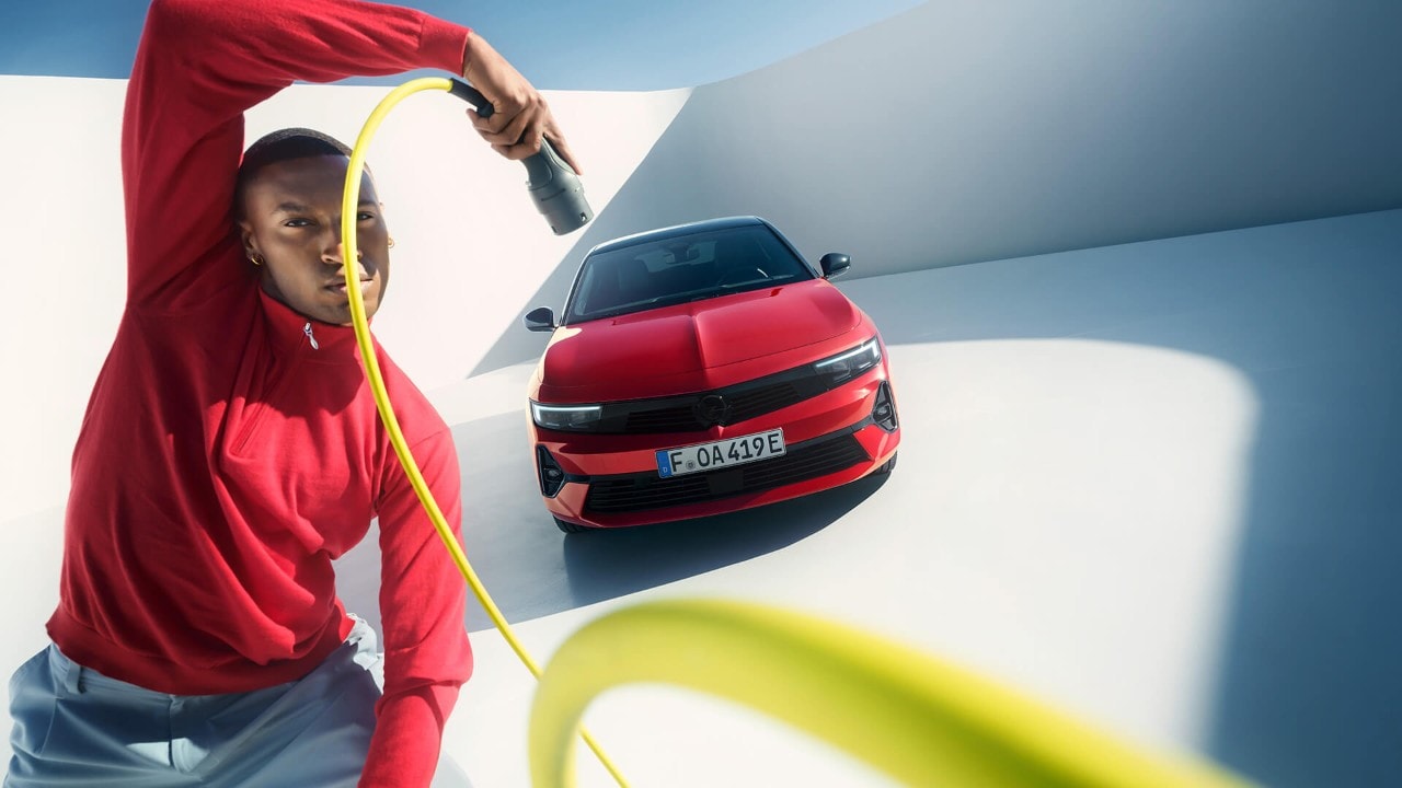 Mann som står foran en rød Opel Astra Sports Tourer med sort tak og holder en ladekabel i hånden